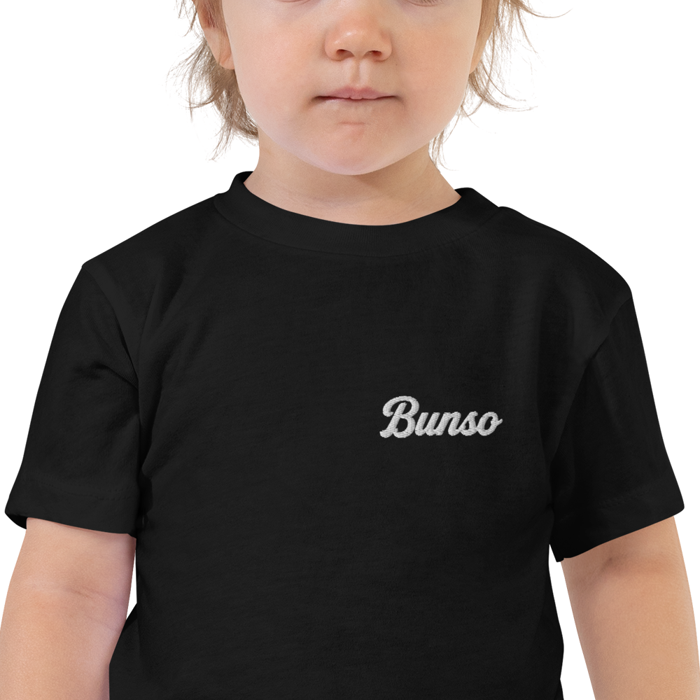 Toddler wearing a black Bunso (Youngest) toddler t-shirt, close-up of the white embroidered name on the left chest