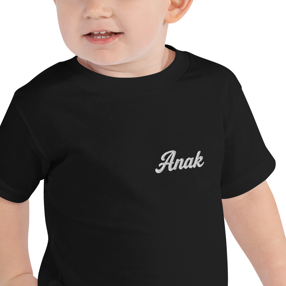 Toddler wearing a black Anak (Child) toddler t-shirt with white embroidered name on the left chest, close-up