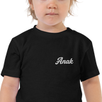 Toddler wearing a black Anak (Child) toddler t-shirt, close-up of the white embroidered name on the left chest